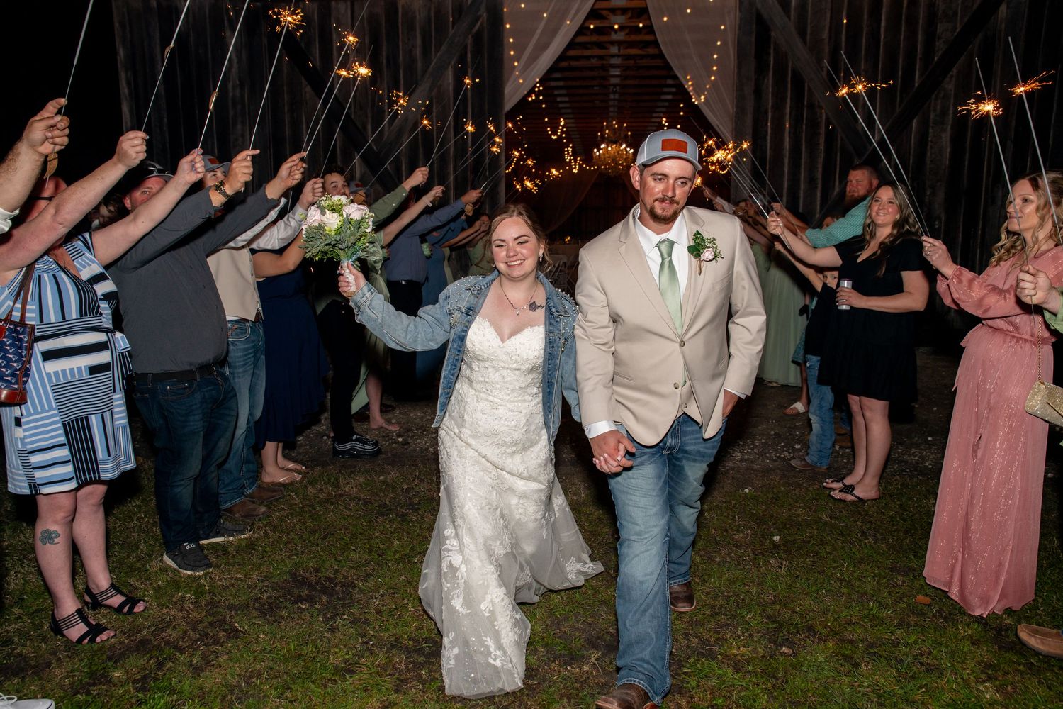 Bride and groom exit barn wedding venue through sparkler tunnel with cheering guests at night.