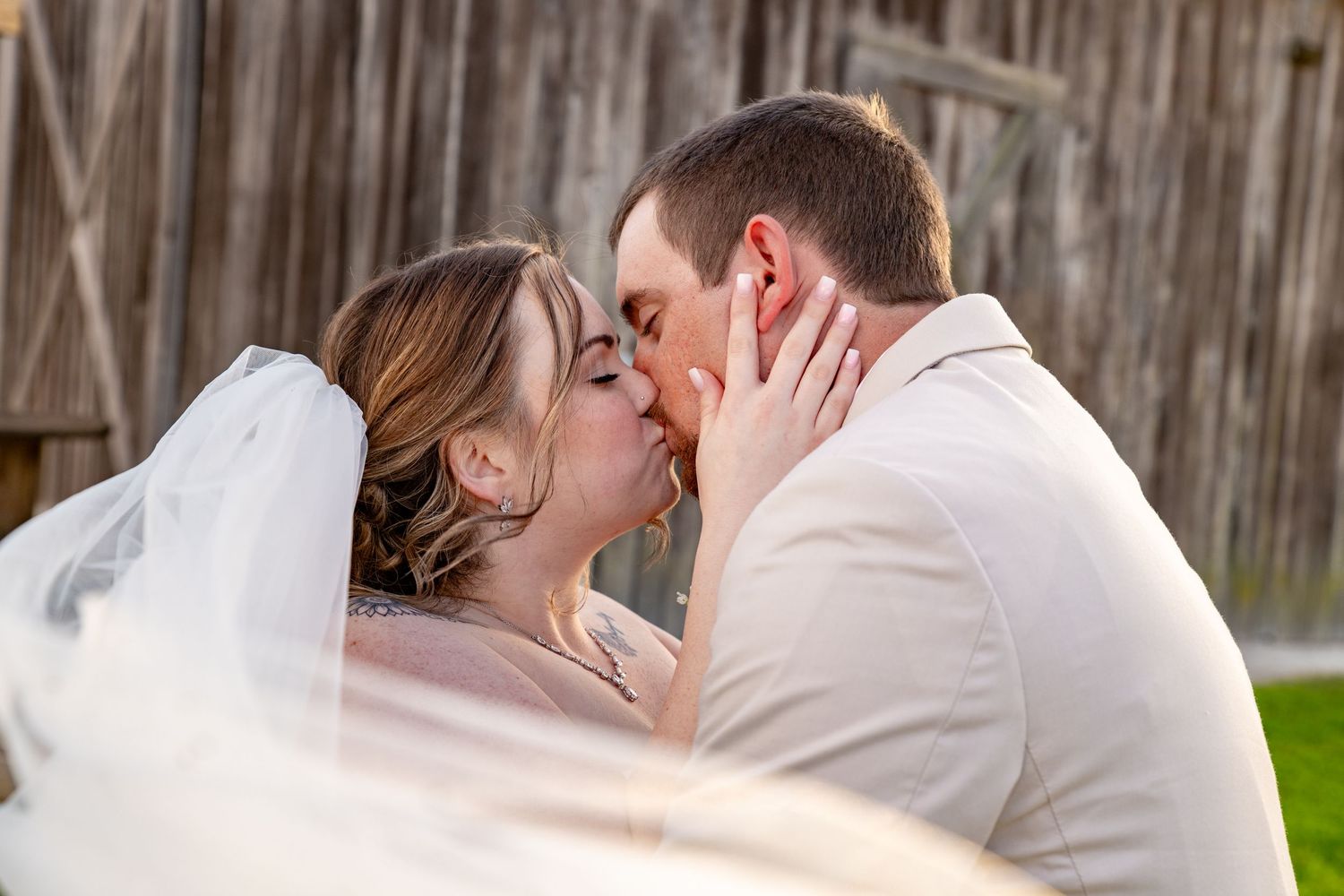 Bride and groom share a romantic kiss under a flowing veil with a rustic wooden barn backdrop.