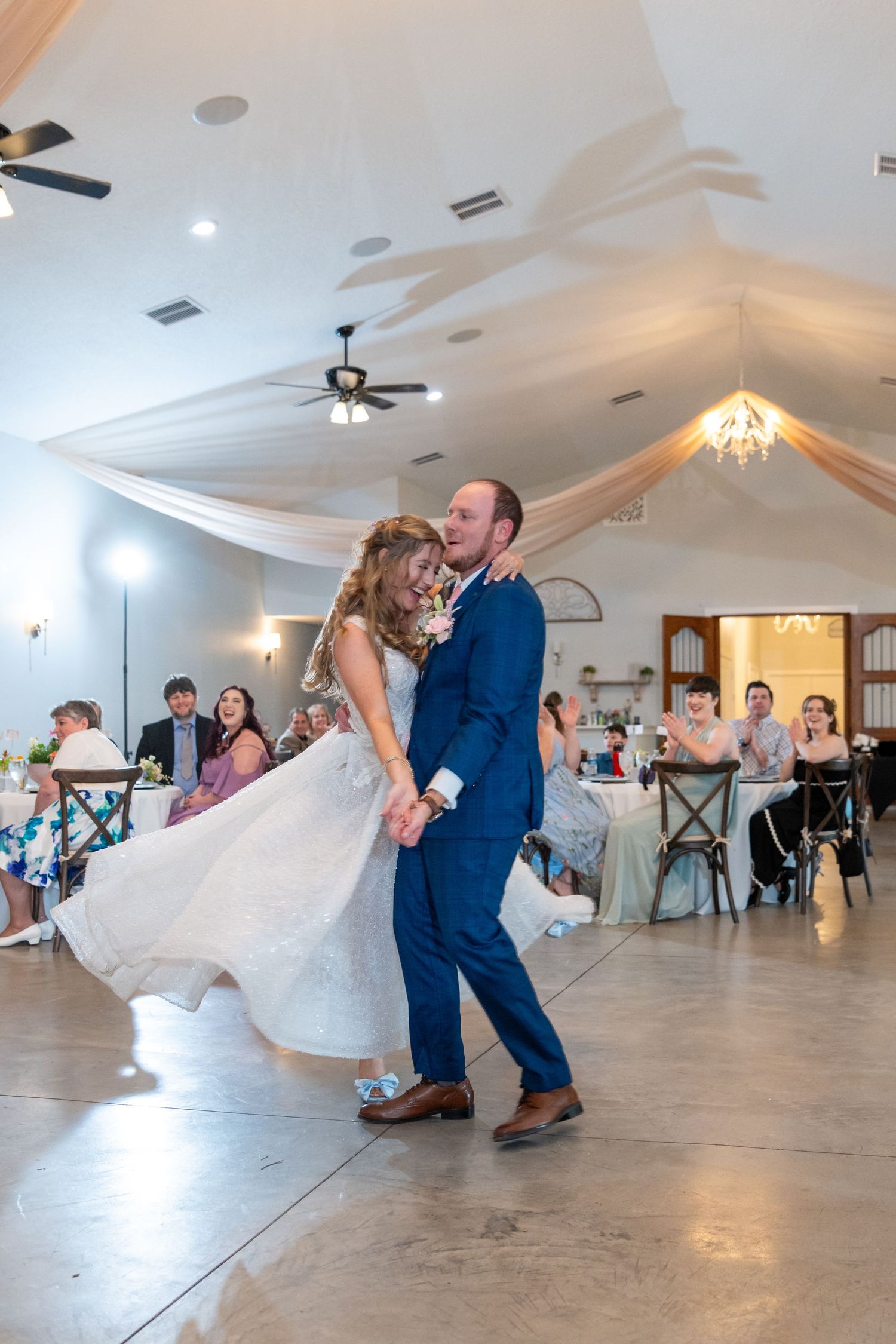 Bride and groom dance joyfully as guests watch in a decorated reception hall with draped ceiling.
