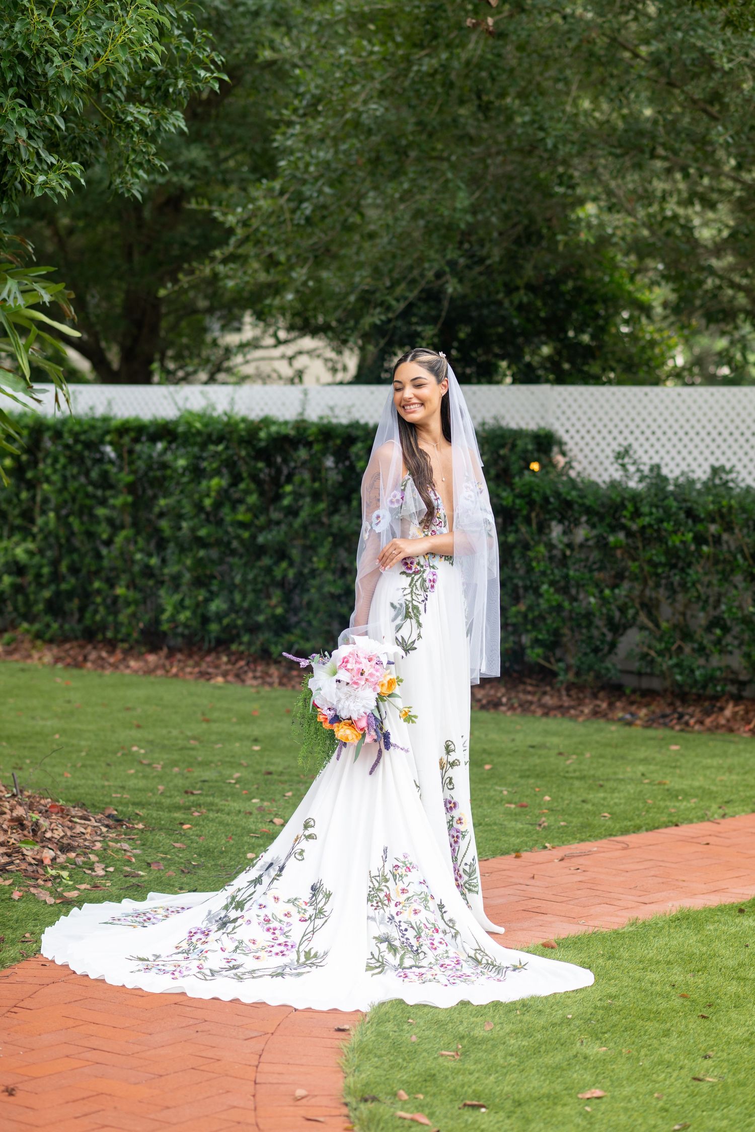 Bride in floral embroidered white wedding gown with veil holding colorful bouquet in garden setting.