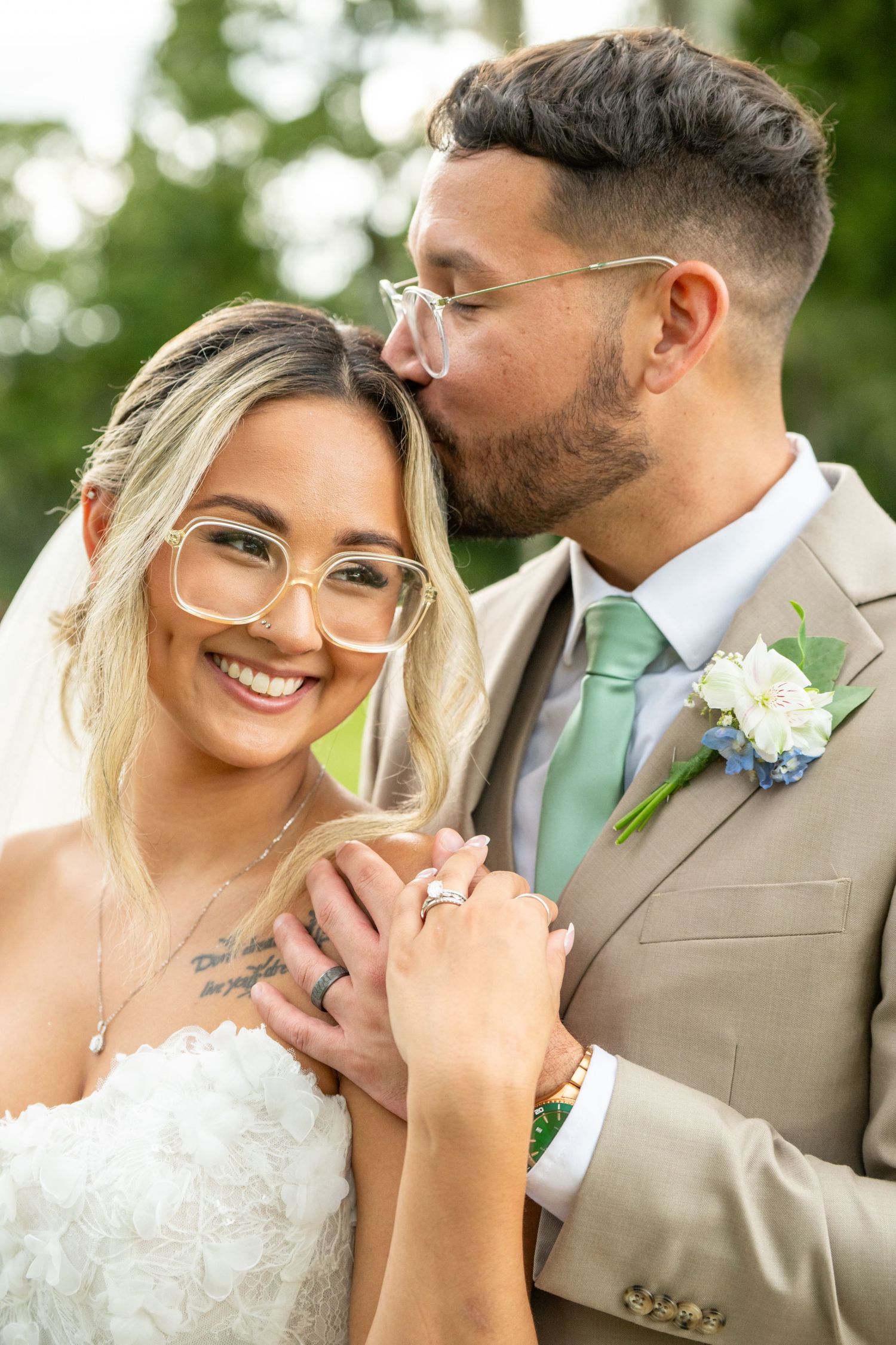 Smiling bride with glasses and groom in tan suit sharing a tender kiss on their wedding day outdoors.