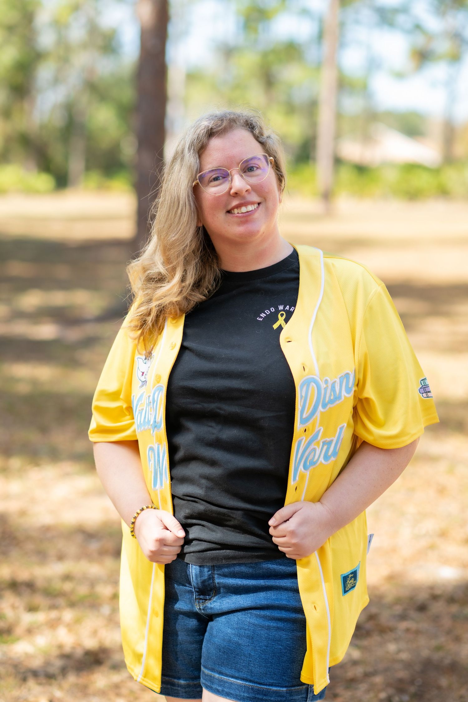 Woman smiling outdoors in yellow Disney World baseball jersey over black tee and jeans, surrounded by pine trees.