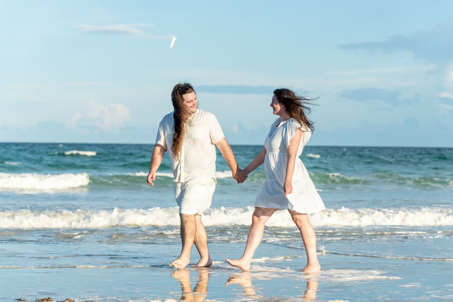 Couple holding hands walking barefoot on a sunny beach with ocean waves, wearing matching white outfits.