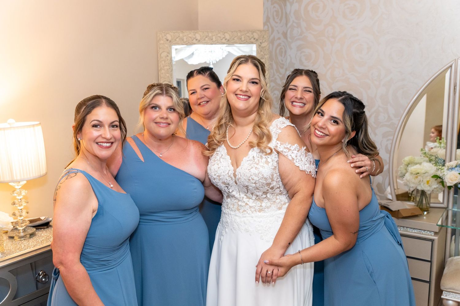 Bride in white lace gown smiles with four bridesmaids in blue dresses in a bright dressing room.