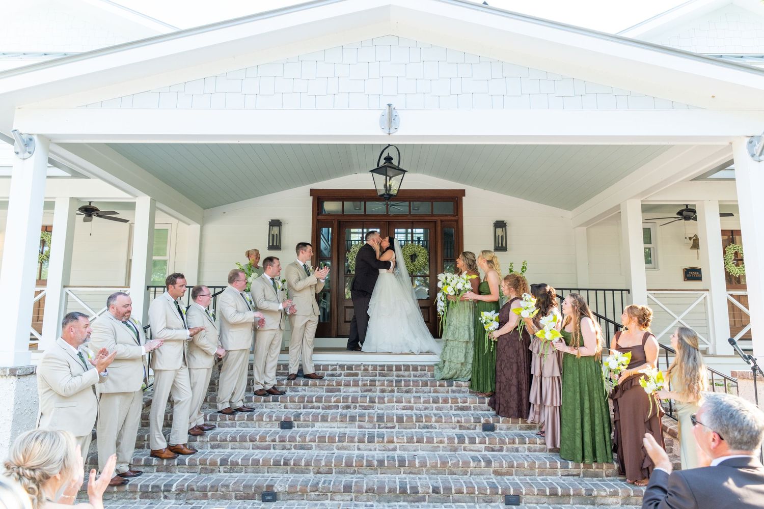Wedding couple kisses on church steps surrounded by groomsmen in tan suits and bridesmaids in green dresses.