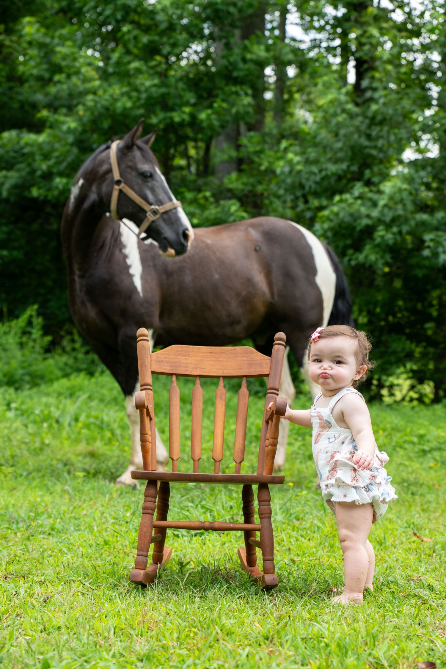A baby girl in a floral outfit stands by a wooden rocking chair on green grass with a black and white horse behind her.