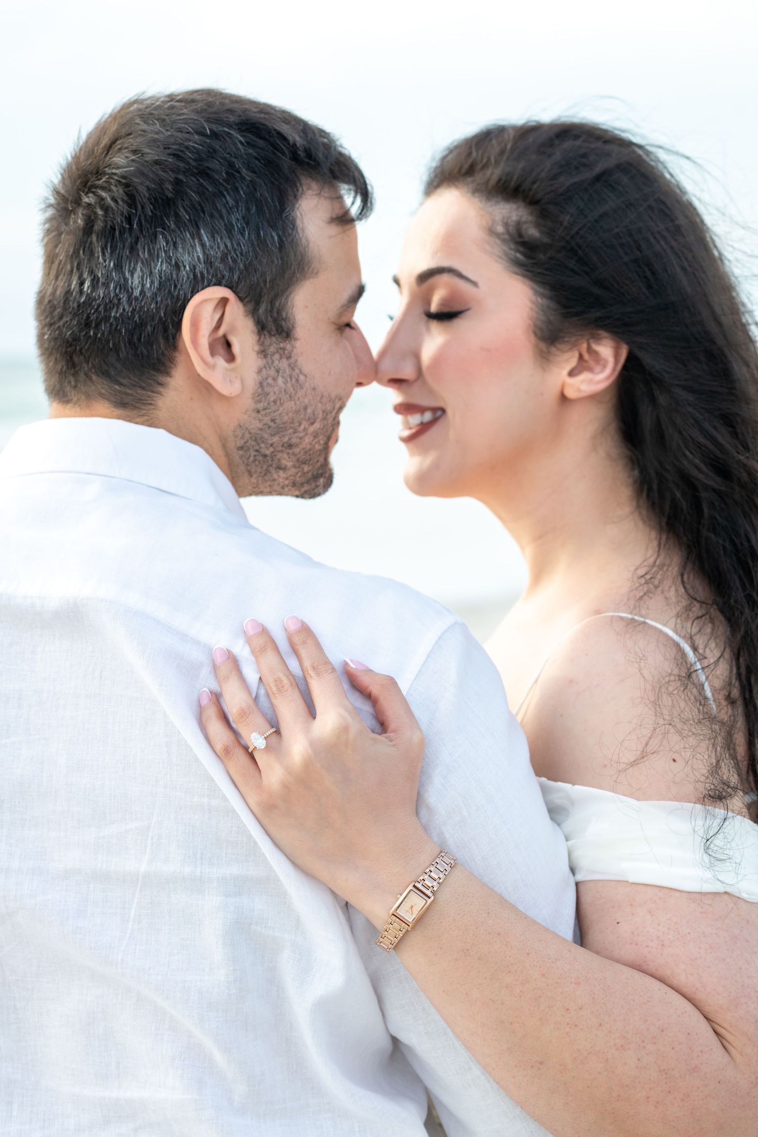 Romantic couple sharing an intimate moment, woman in white dress with ring and gold watch, man in white shirt outdoors.