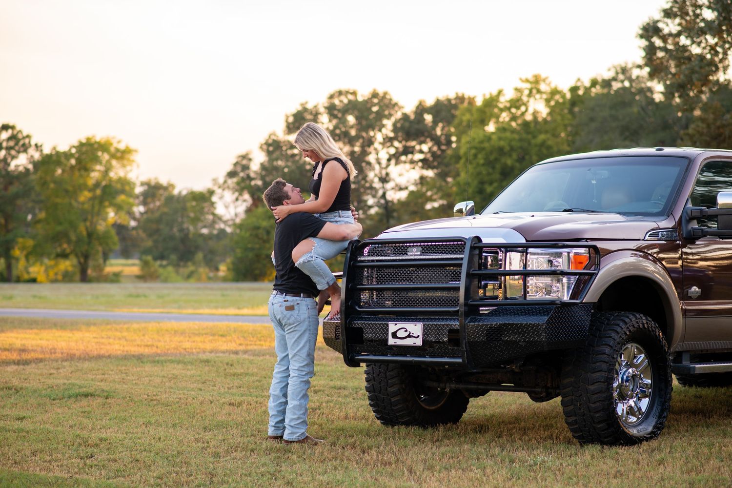 Man lifting woman in front of a large dark pickup truck at sunset in an open grassy field.