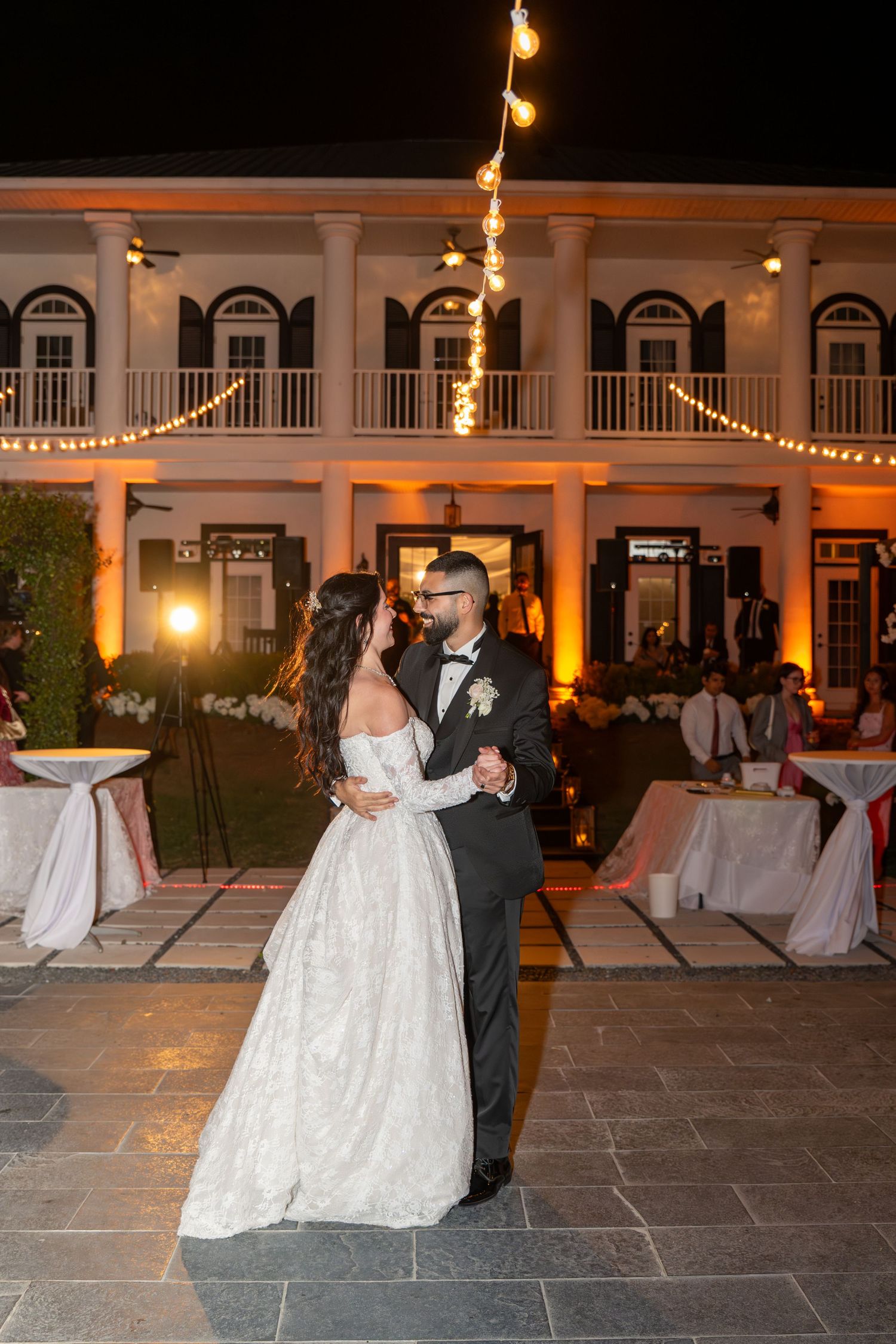 Bride and groom share their first dance outside an elegant venue lit with warm string lights at night.