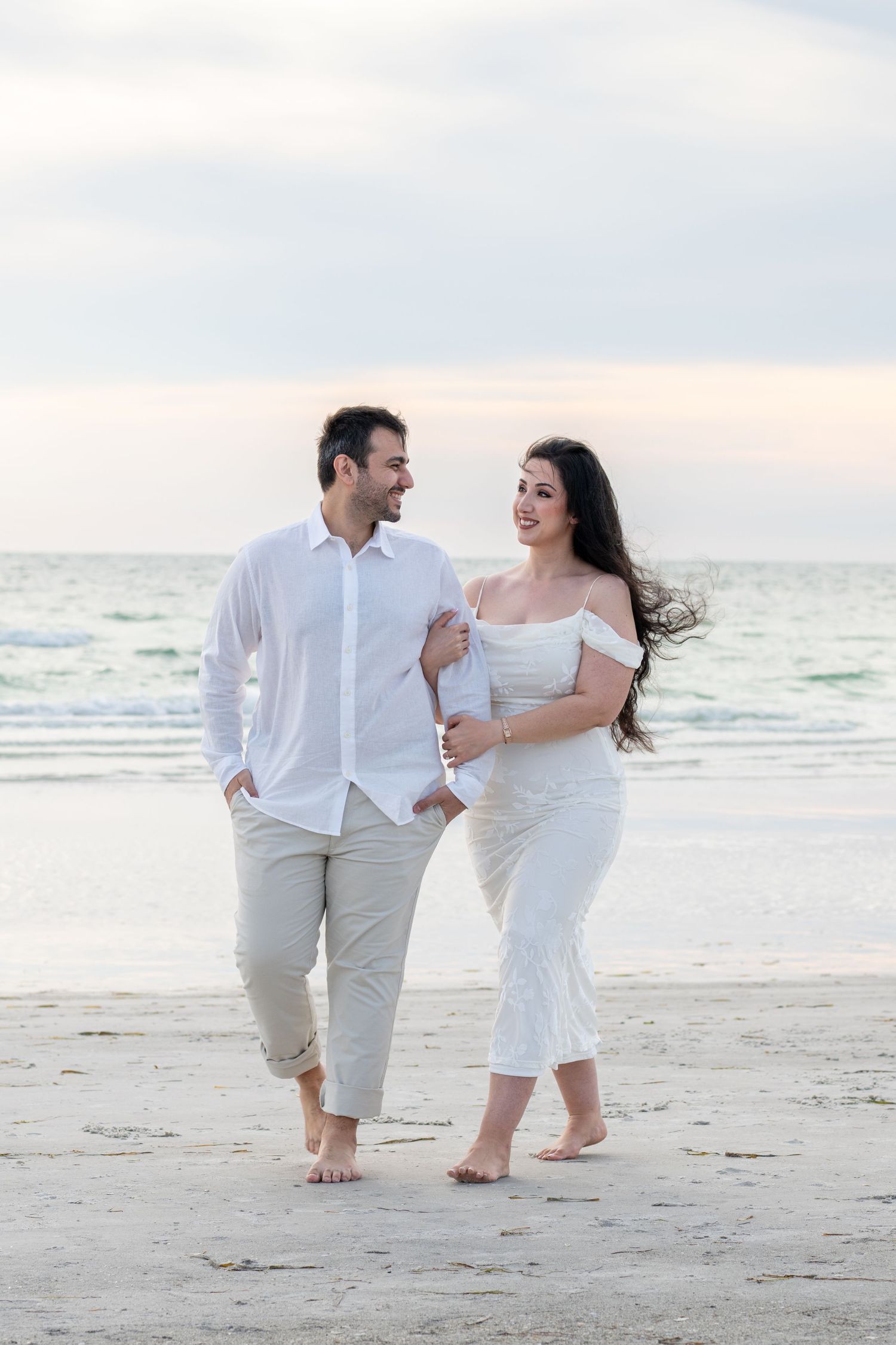 Couple in white outfits walking barefoot on a sandy beach at sunset, smiling at each other near the ocean.