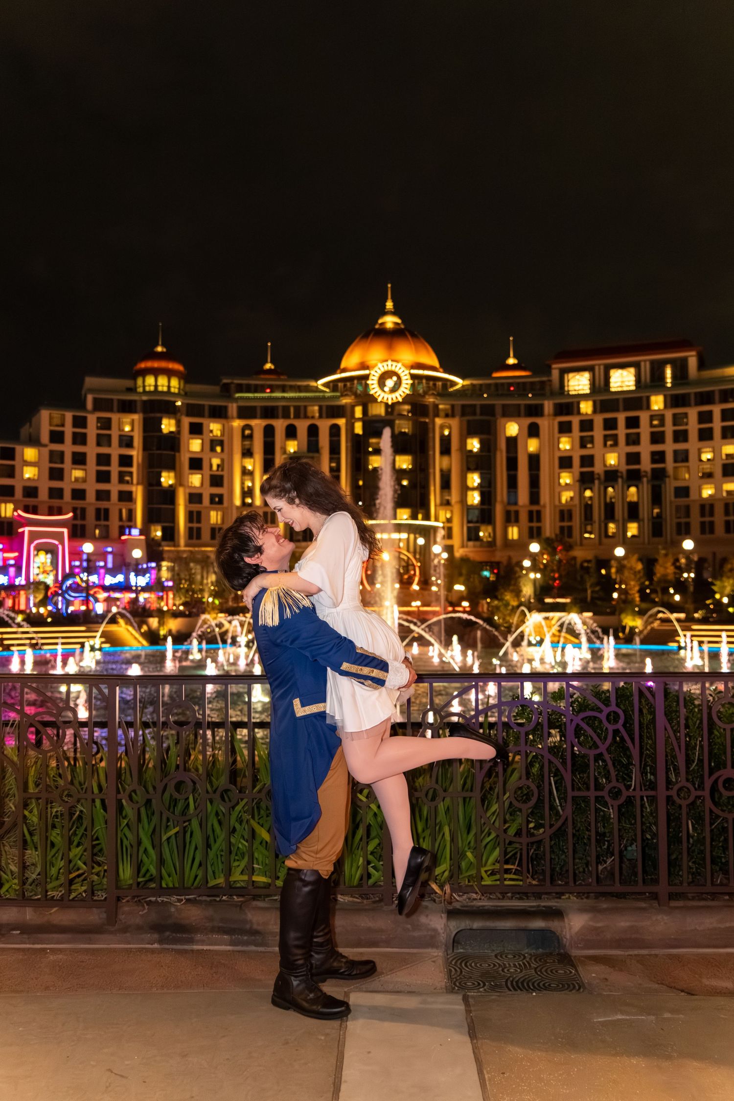 Couple embracing at night in front of illuminated hotel with colorful fountains and golden dome.