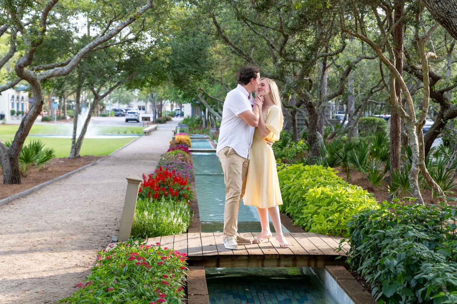 Couple sharing a romantic kiss on a wooden bridge surrounded by lush gardens and red flowers in a scenic park.