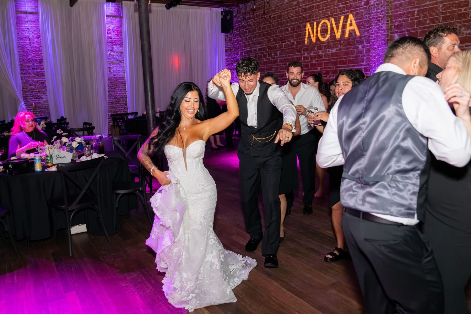 Bride and groom dance joyfully at wedding reception with guests, purple lighting, and NOVA sign on brick wall.