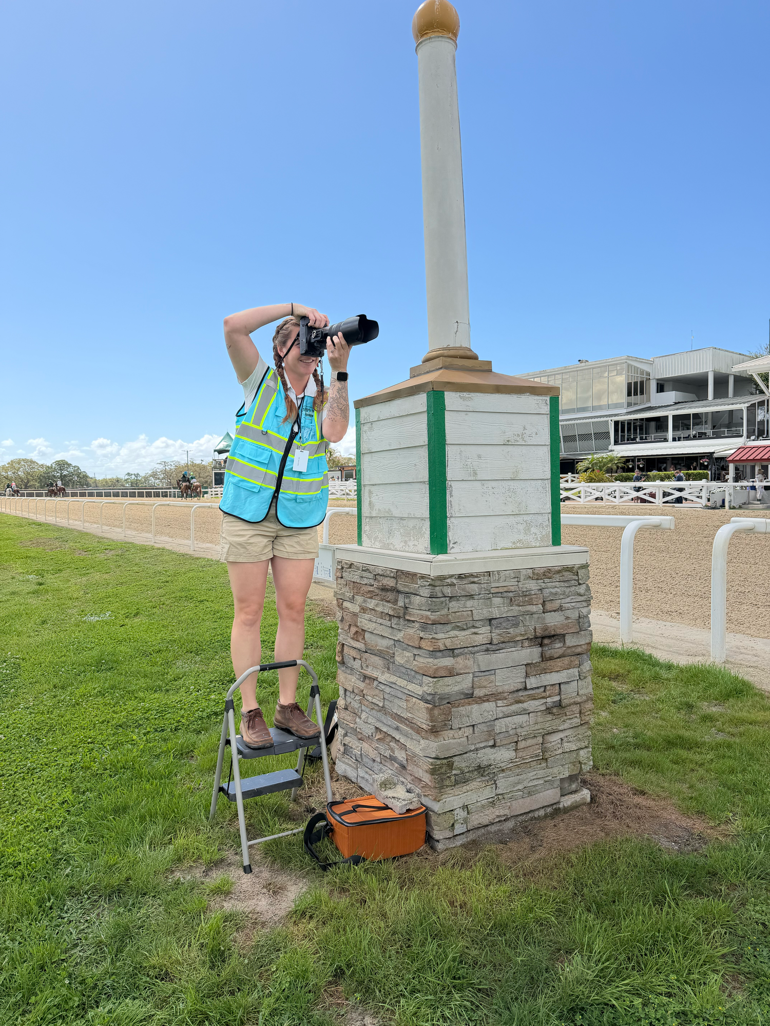 Photographer in safety vest stands on ladder at stone monument pillar at a racetrack, capturing images on a sunny day.