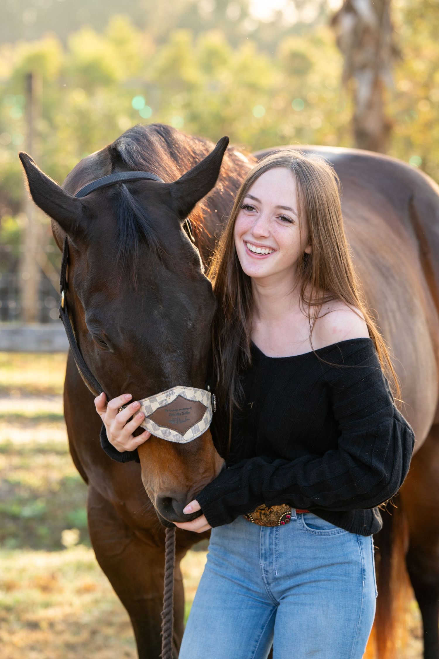 Smiling teenage girl with long brown hair posing with chestnut horse outdoors.