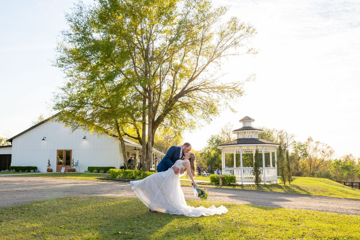 Groom dips bride near white chapel and gazebo on lush green lawn in golden afternoon light.