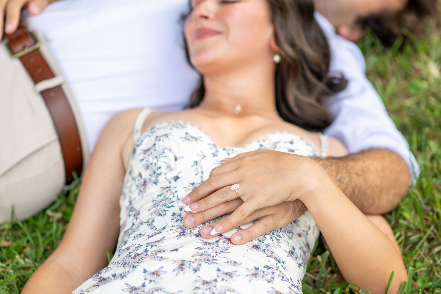 Couple lying on grass holding hands, woman wearing a floral dress with an engagement ring visible.