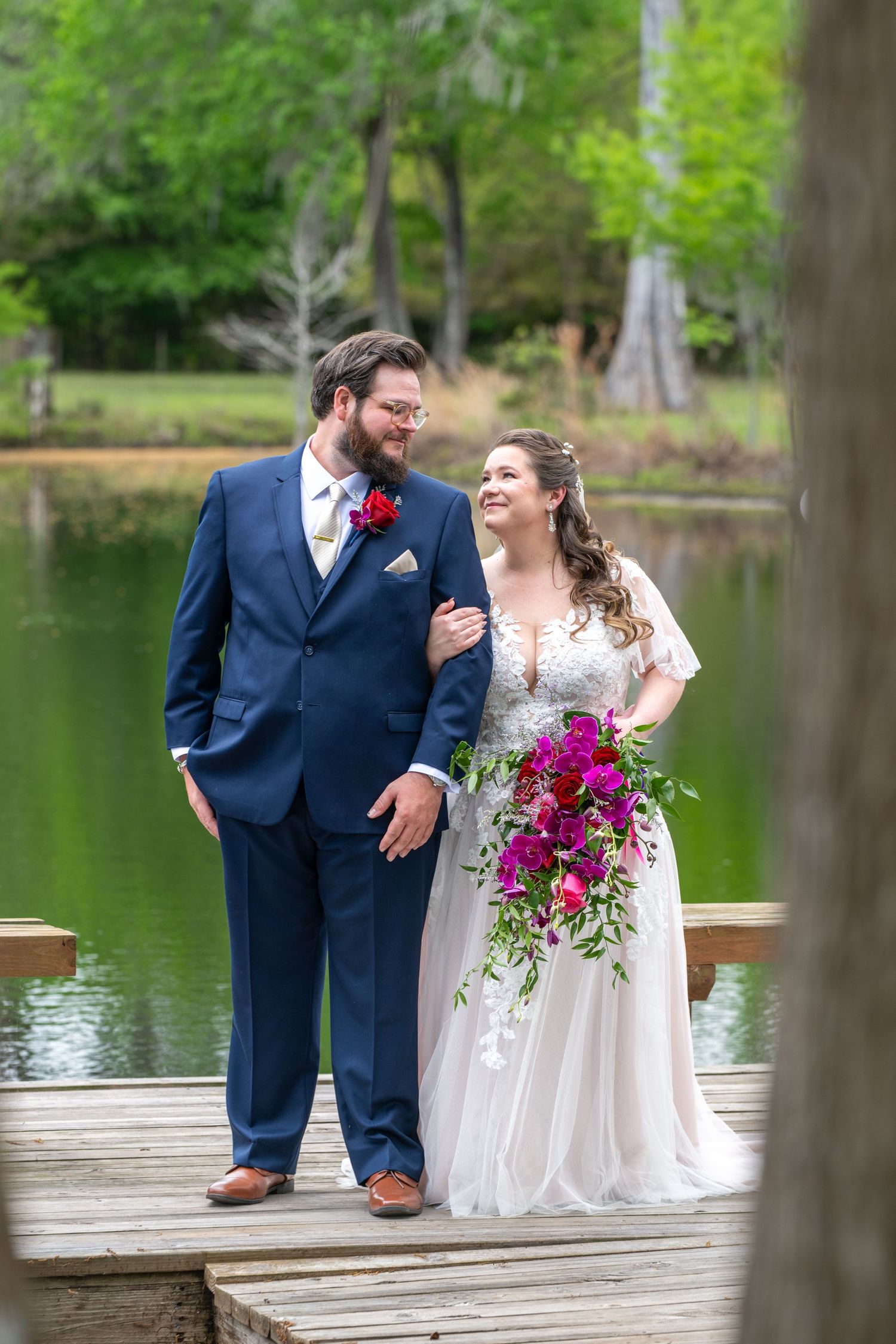 Smiling bride holds vibrant pink bouquet beside bearded groom in navy suit on a lakeside wooden dock.