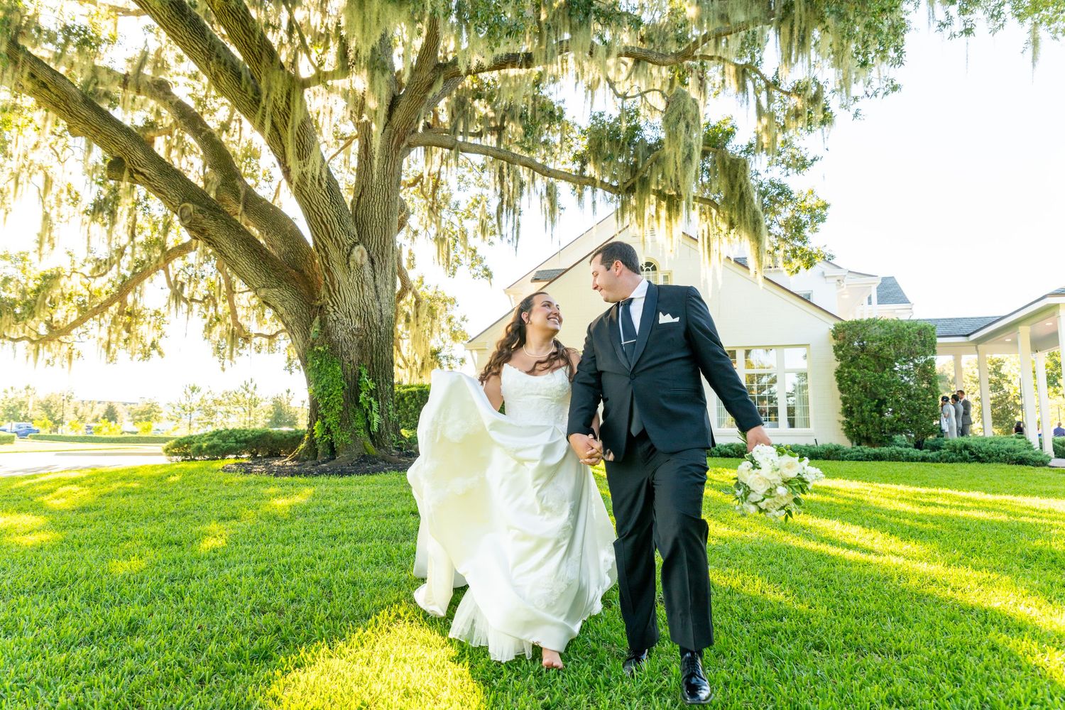 Bride and groom walk hand-in-hand on lush green lawn beneath a sprawling oak tree draped in Spanish moss.