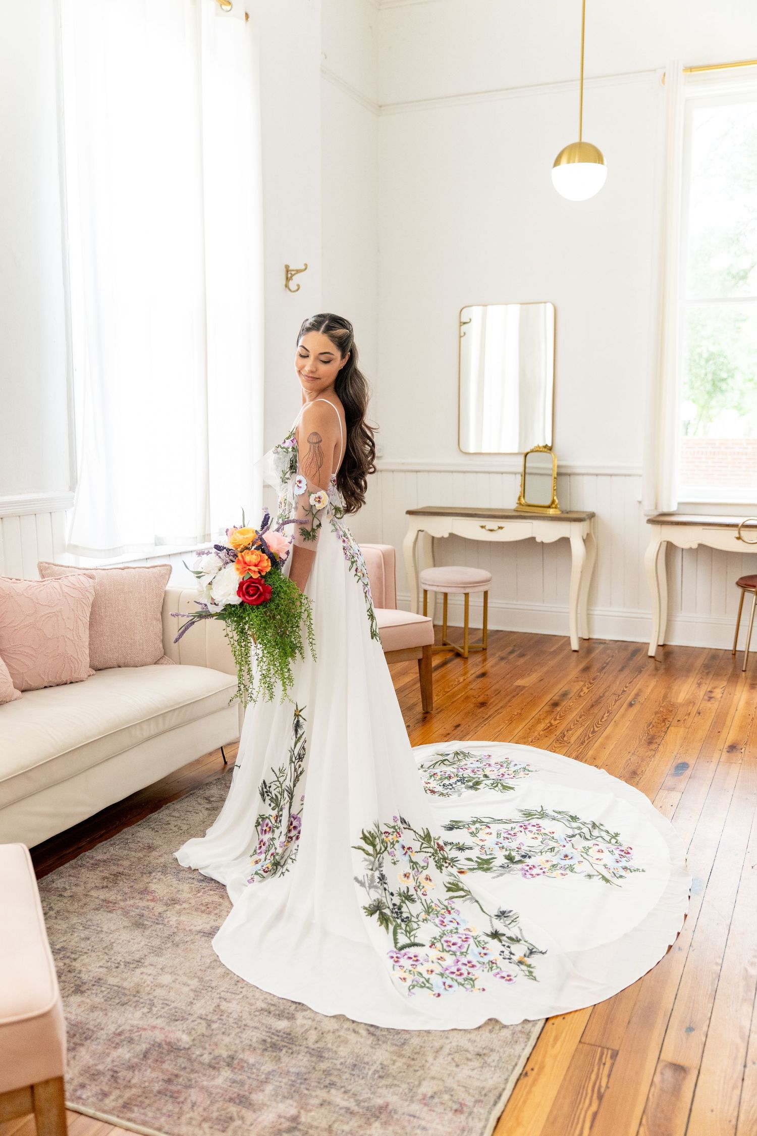 Bride in floral-embroidered white wedding gown with long train holds colorful bouquet in bright elegant bridal suite.