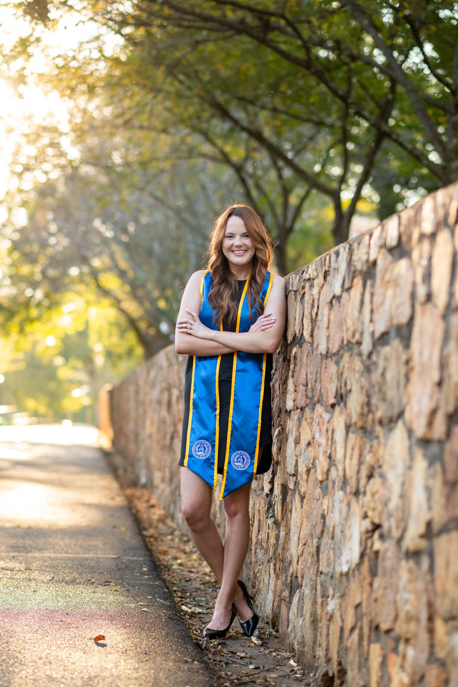 Smiling graduate in blue stole leans against stone wall on tree-lined campus path.