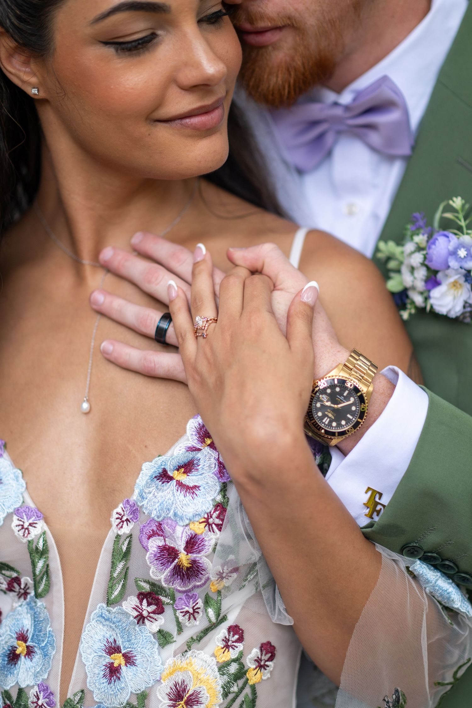Bride and groom holding hands showing wedding rings, bride in floral embroidered gown, groom in green suit with lavender bow tie.