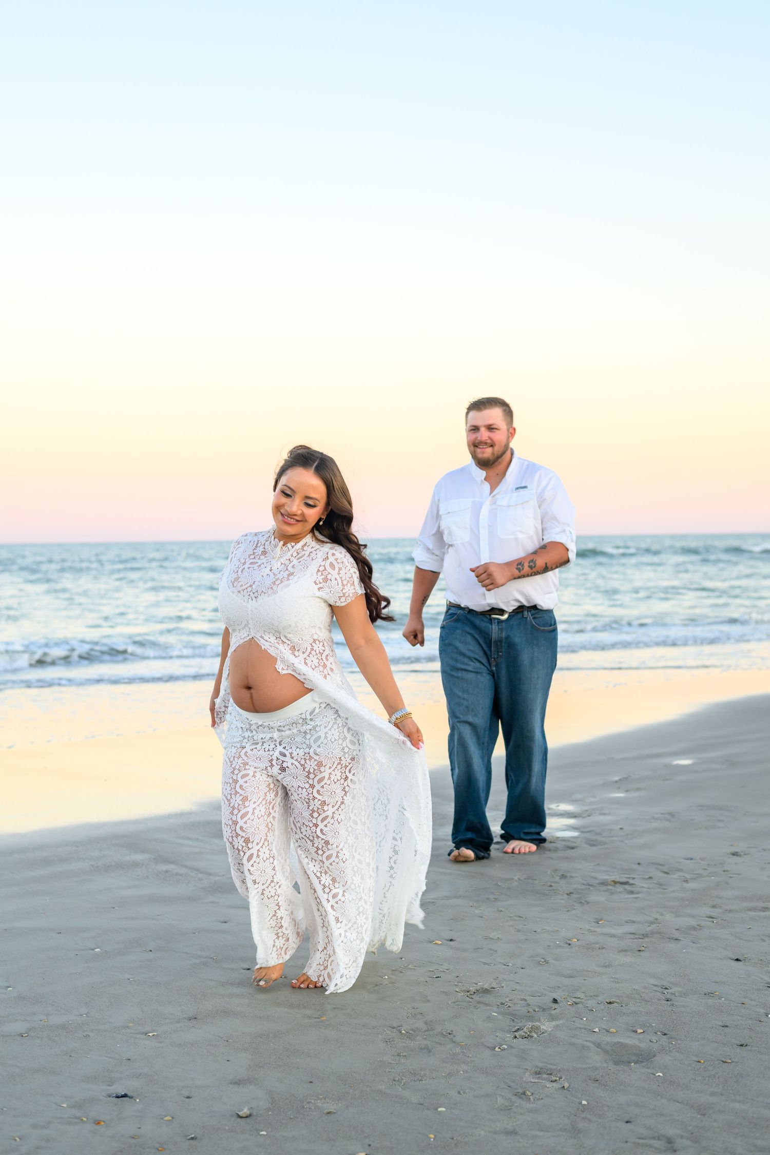 Pregnant woman in a white lace dress walks along the beach at sunset while a smiling man in jeans follows behind her.