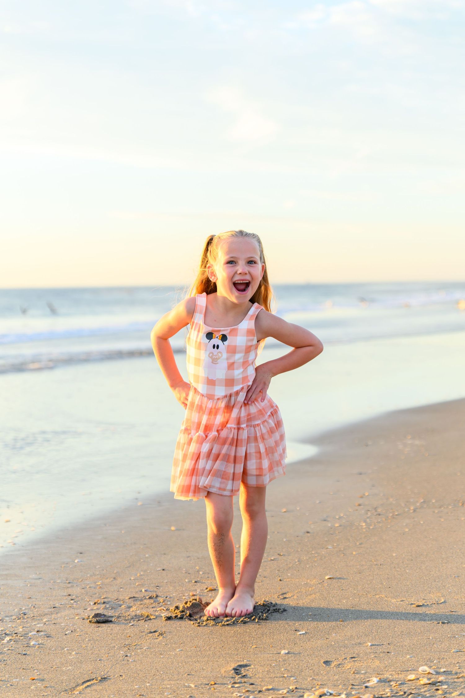 Smiling young girl in a pink plaid dress with pigtails stands barefoot on a sandy beach at sunset with gentle ocean waves behind her.