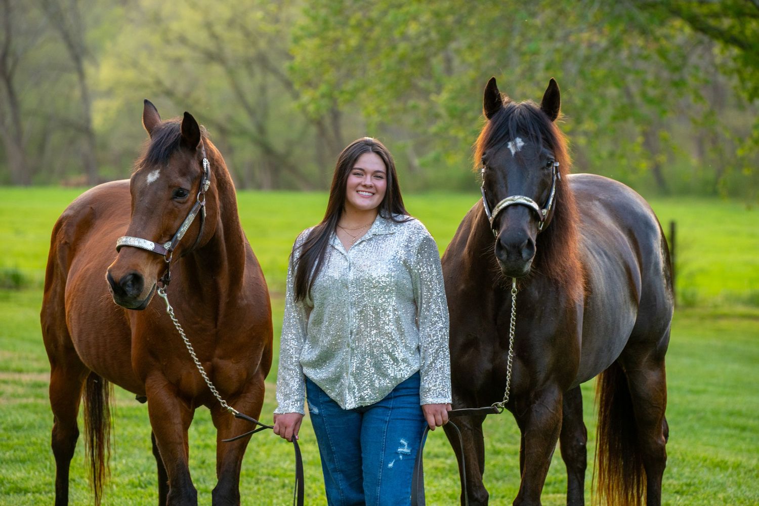 Young woman smiling between two bay horses on green grass, holding lead ropes outdoors.