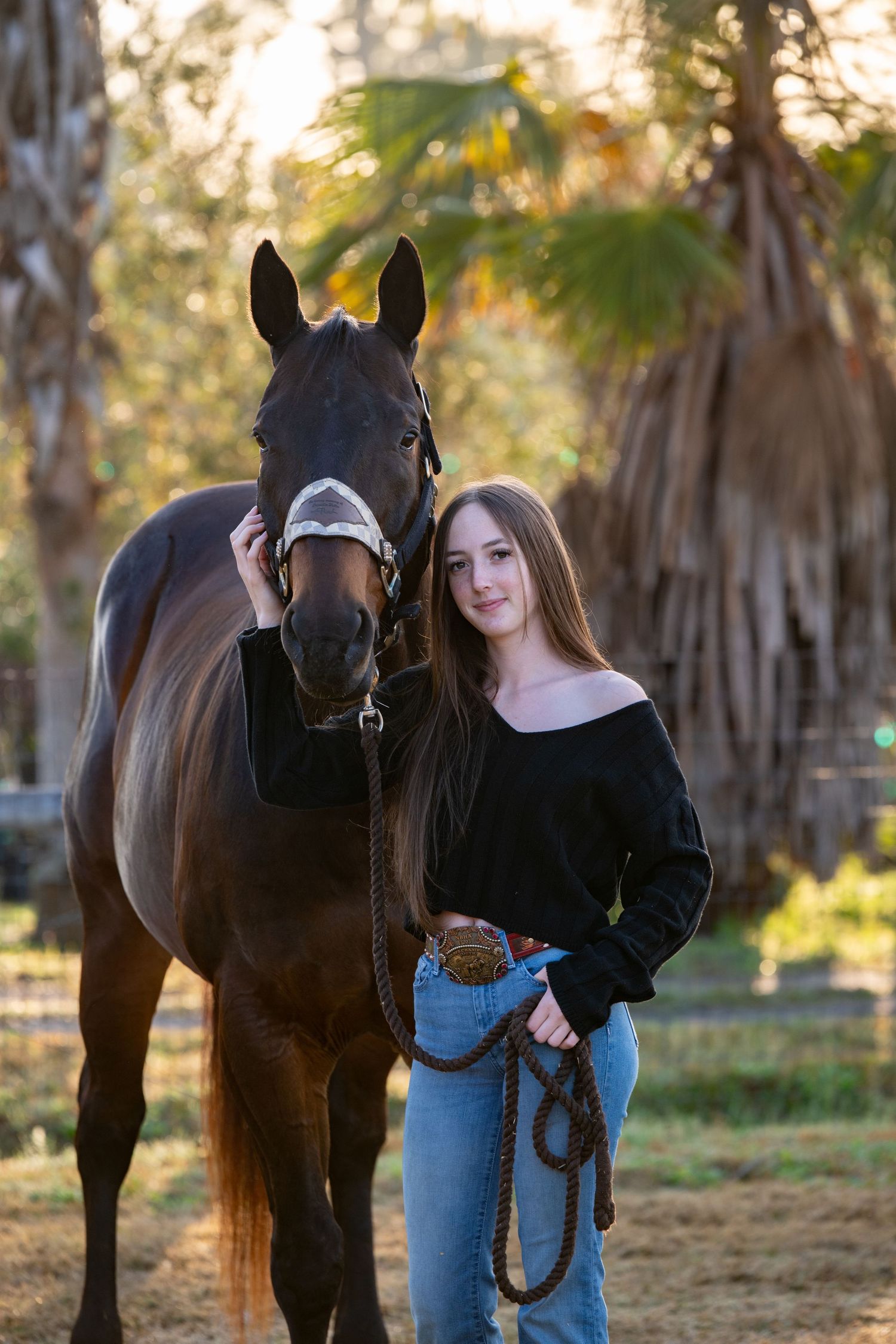 Young woman in black off-shoulder top and jeans stands beside a dark brown horse outdoors near palm trees.