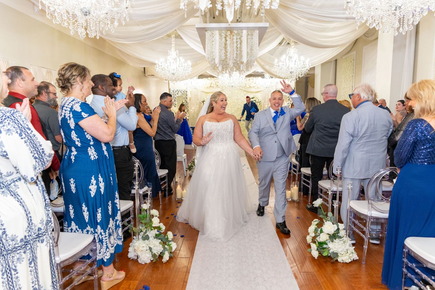 Bride and groom walk down the aisle at an elegant indoor wedding ceremony with chandeliers and guests applauding.