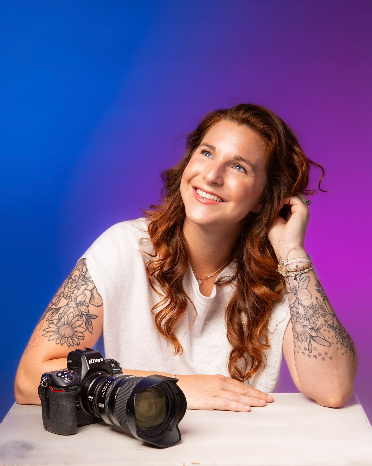 Smiling woman with tattoos and red hair poses with Nikon camera against colorful purple-blue backdrop.