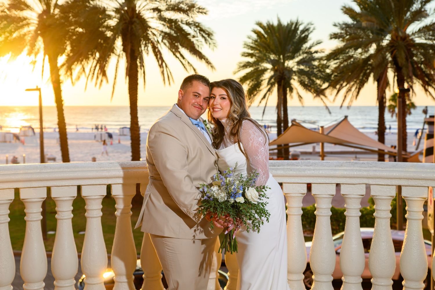 Smiling bride and groom hold a bouquet on a white balustrade terrace overlooking palm trees and a sunset beach.