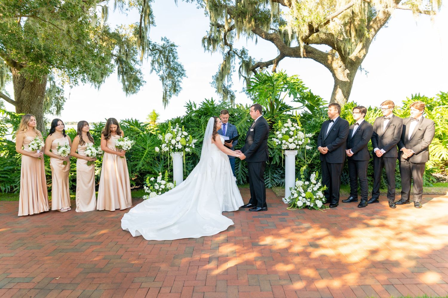 Bride and groom exchange vows outdoors surrounded by bridesmaids in champagne gowns and groomsmen under a mossy oak tree.