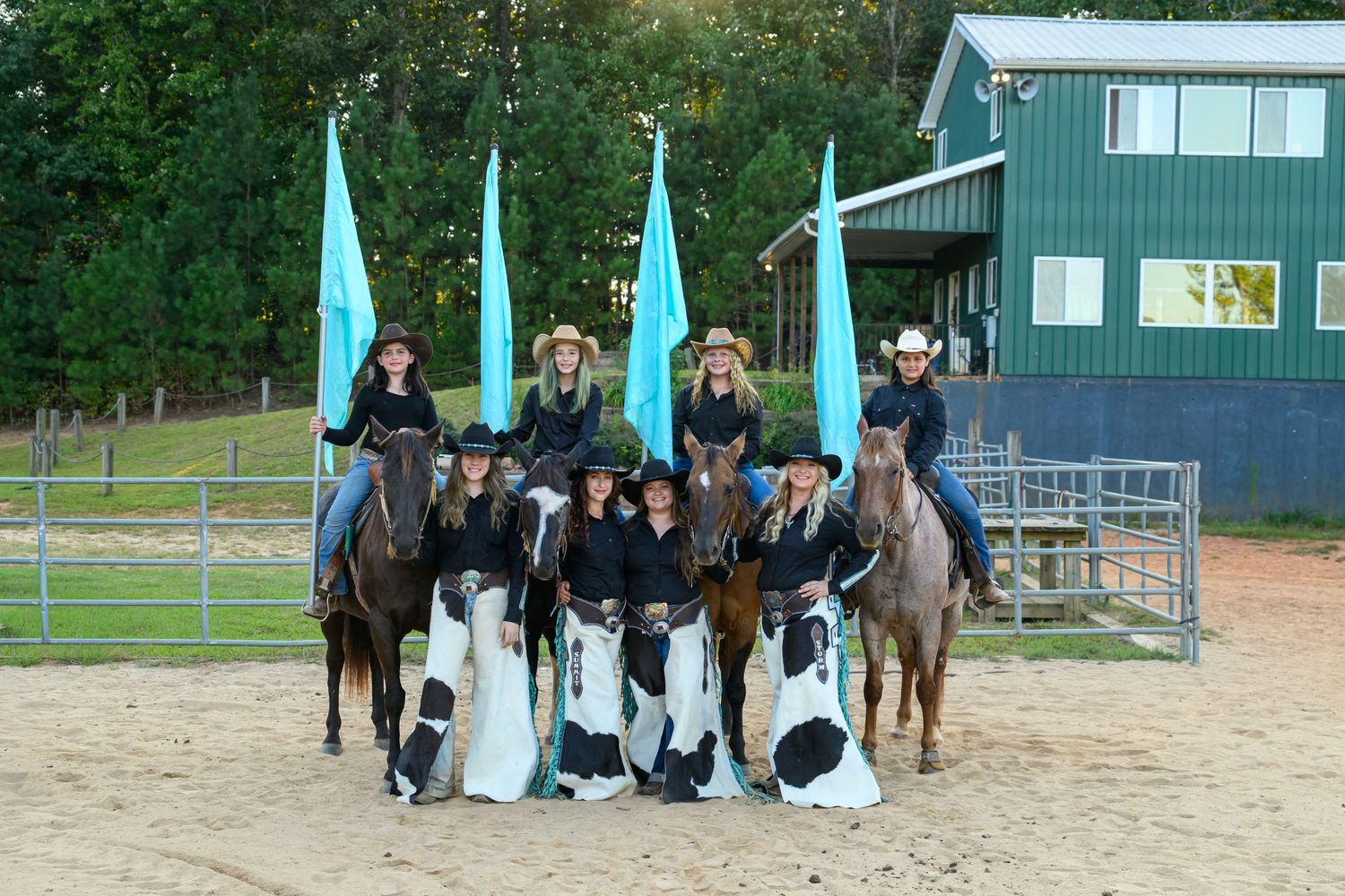 A western equestrian team poses with horses and turquoise flags on a sandy arena near a green barn.