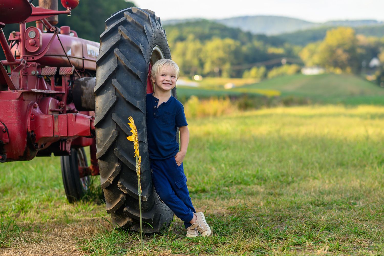 Smiling blonde child in navy jumpsuit leaning against red tractor tire in a green farm field.