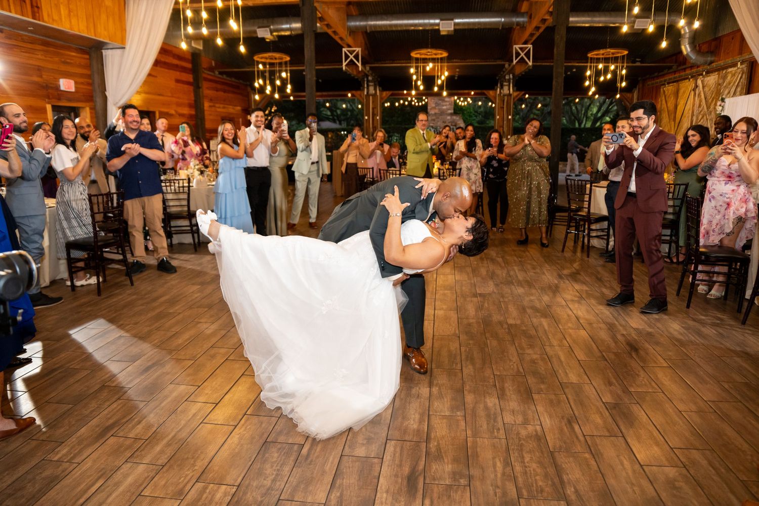 Groom dips bride during first dance at rustic wedding reception with guests applauding in background.