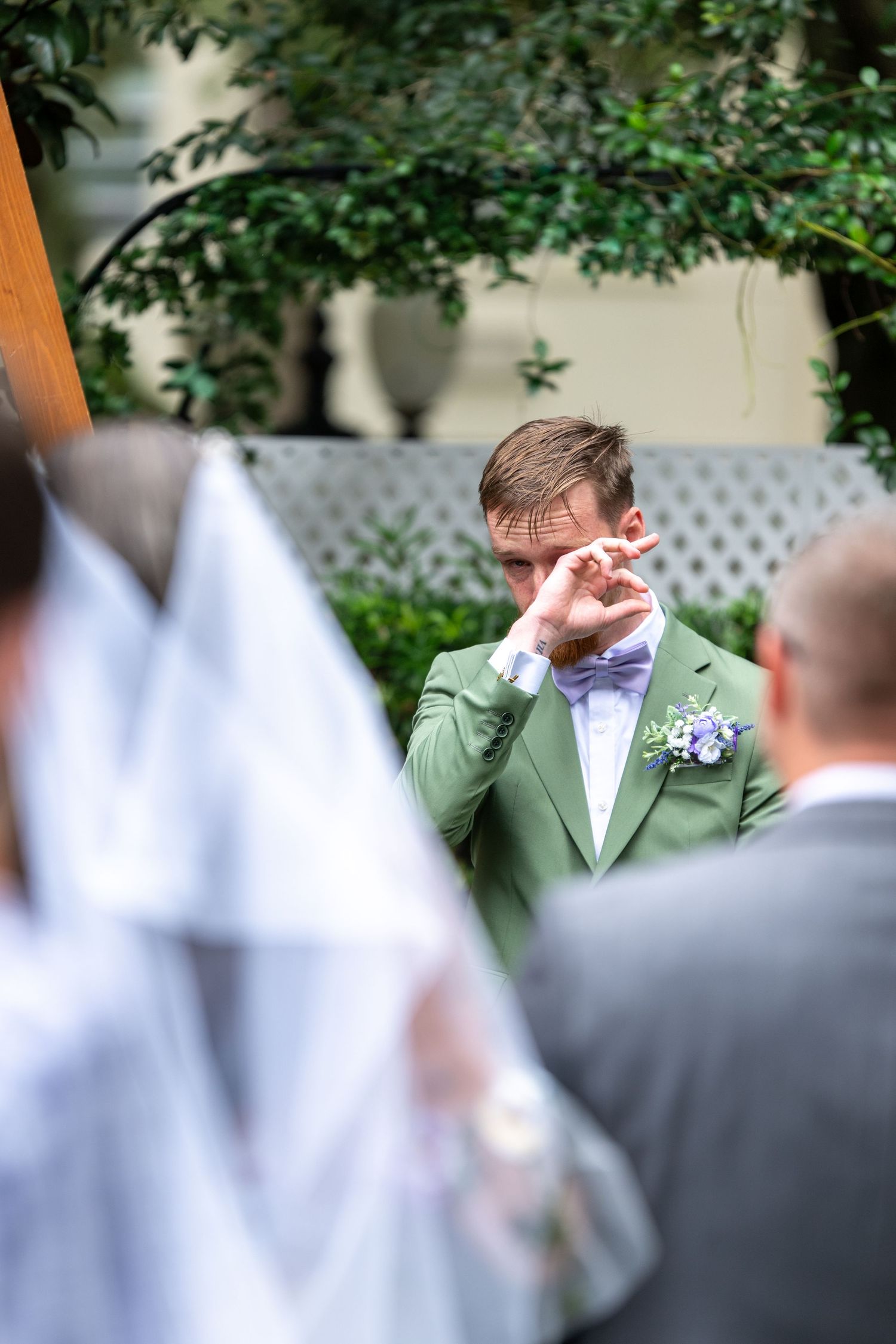 Emotional groom in green suit and bow tie wiping tears as bride approaches in outdoor wedding ceremony.