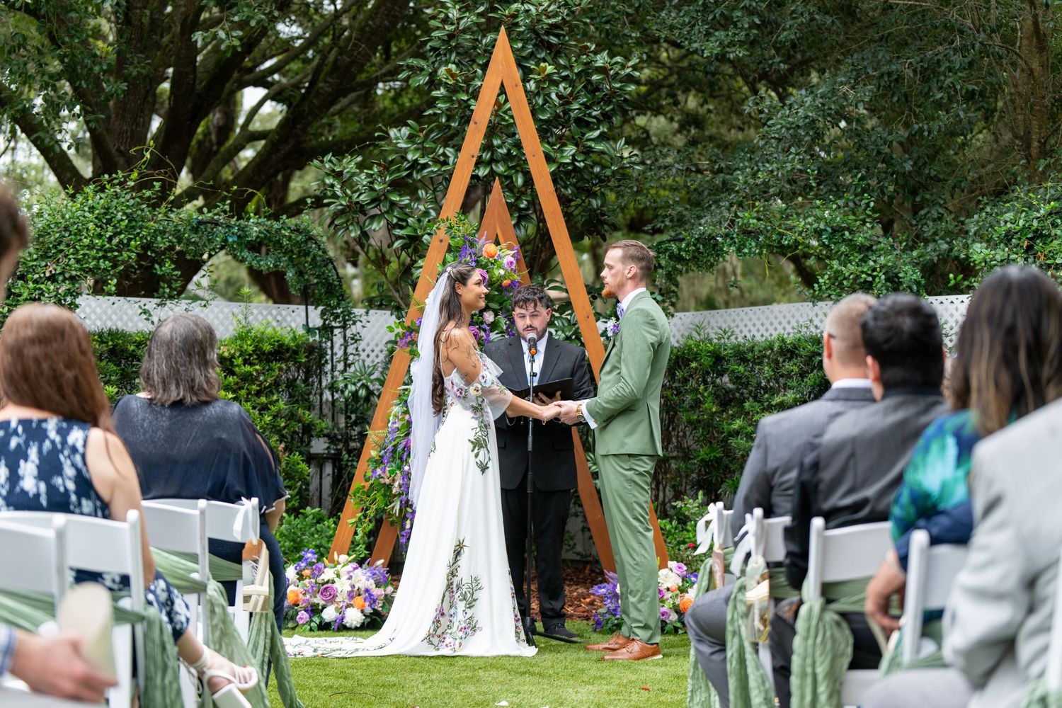 Outdoor wedding ceremony with couple exchanging vows under a wooden triangle arch decorated with flowers and greenery.