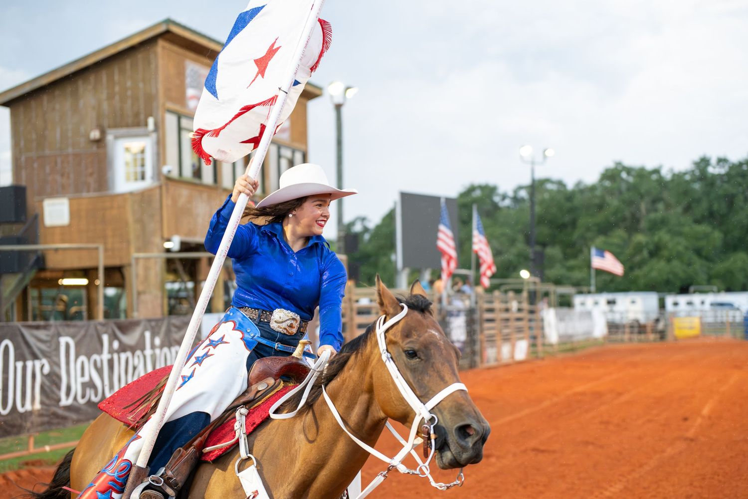 A cowgirl in blue rides a horse carrying flags at a rodeo arena with American flags in the background.