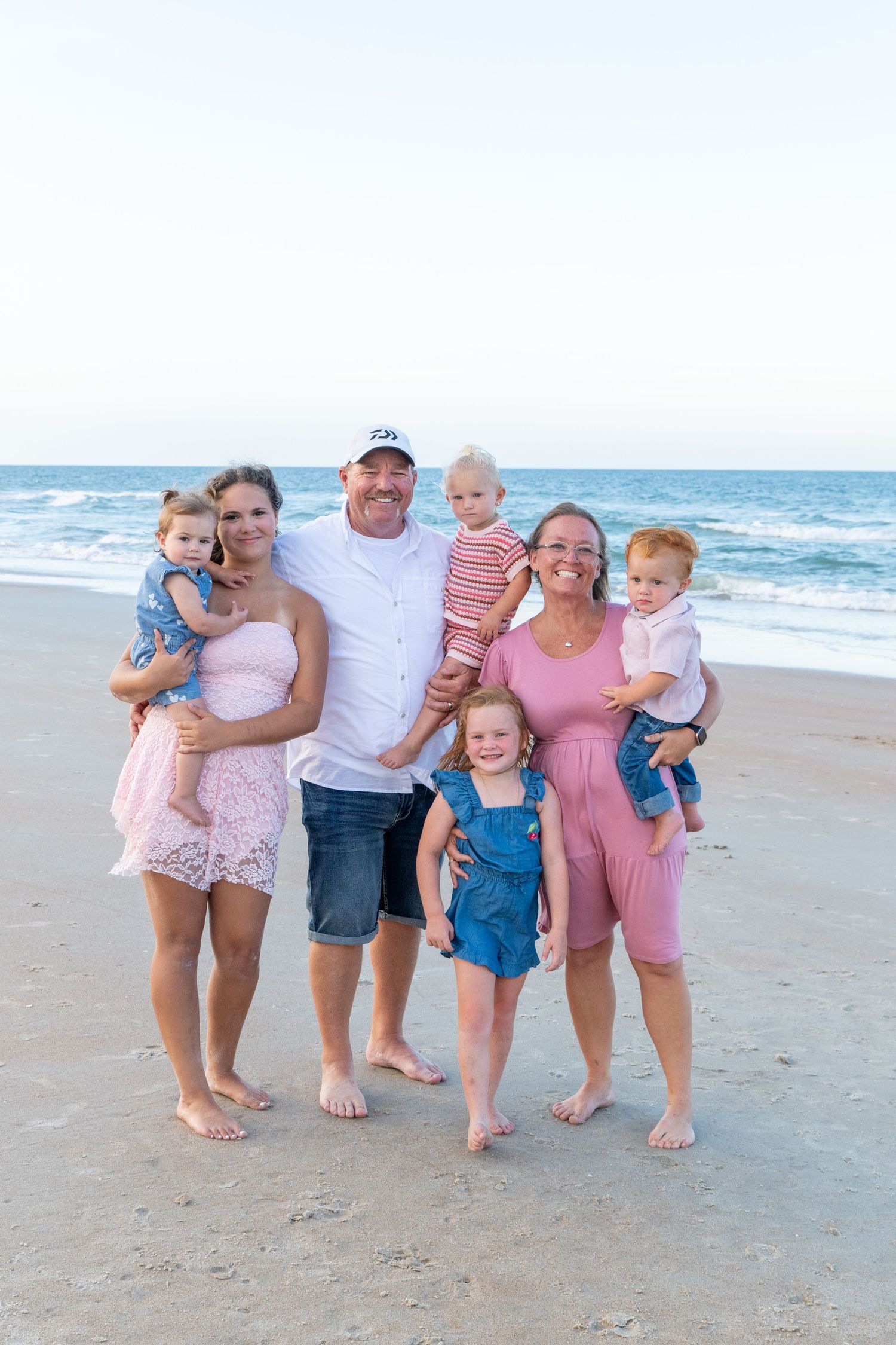 Family of seven smiling together on a sandy beach with ocean waves in the background on a clear day.