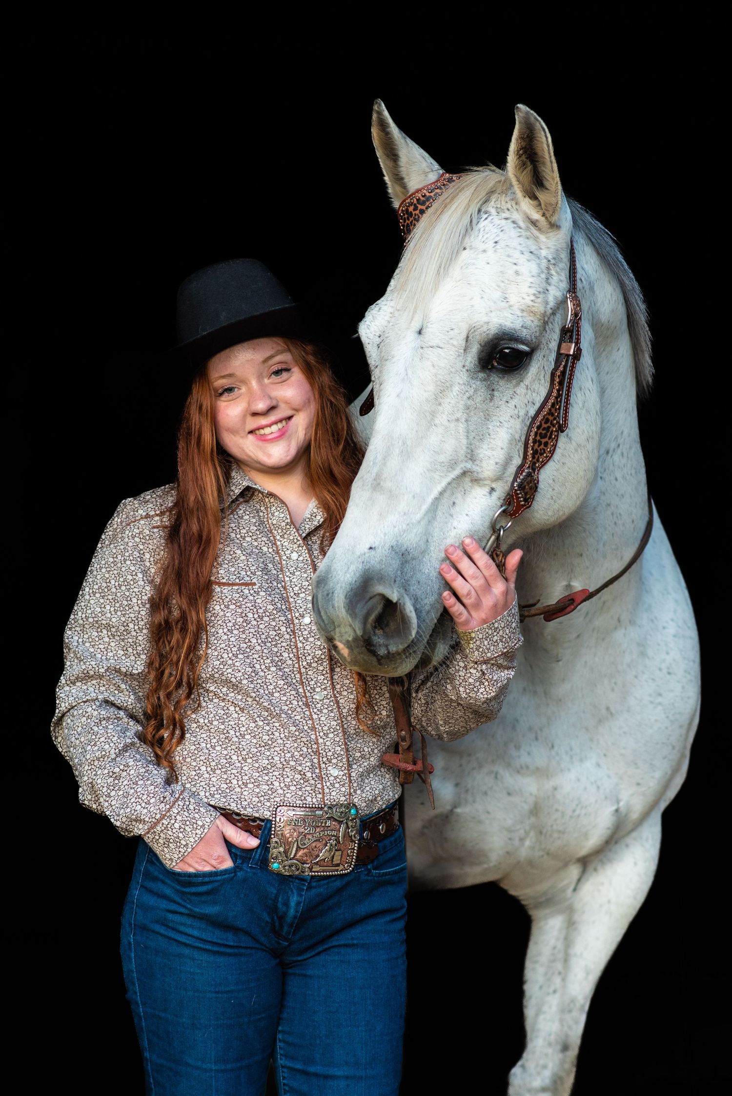 Young cowgirl in black hat and floral shirt smiles beside a white horse against a dark background.
