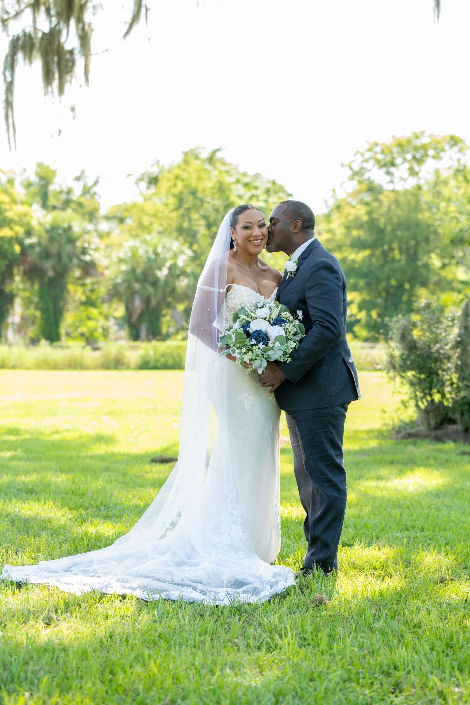 Bride and groom share a romantic kiss outdoors on green grass, bride in white gown with veil holding blue bouquet.
