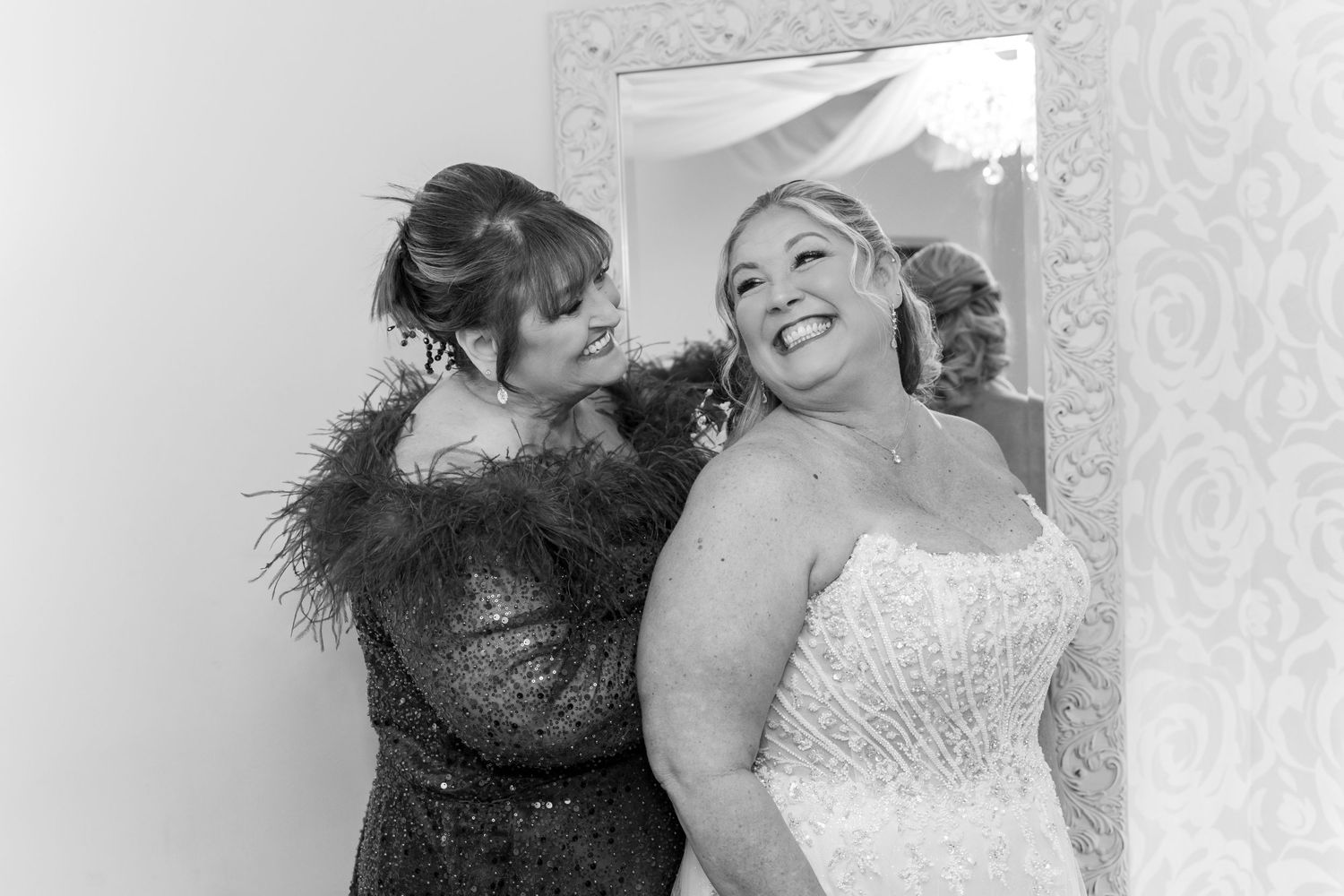 Black and white photo of a bride in a strapless gown laughing with a woman in a feathered dress helping with her earring.