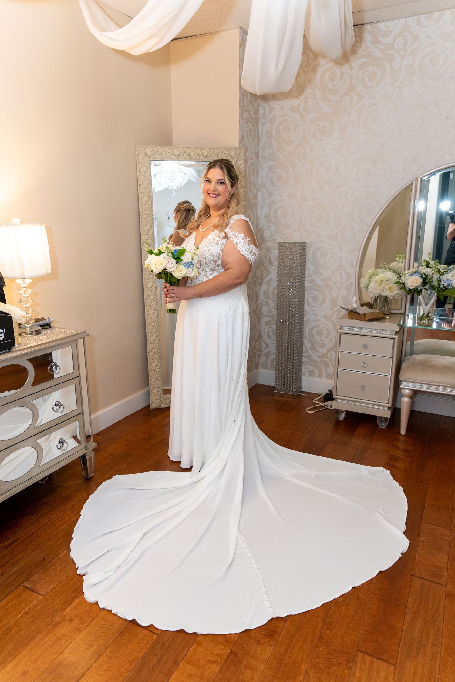 Bride in white lace wedding gown with train holding bouquet, standing in elegant bridal suite with mirror.