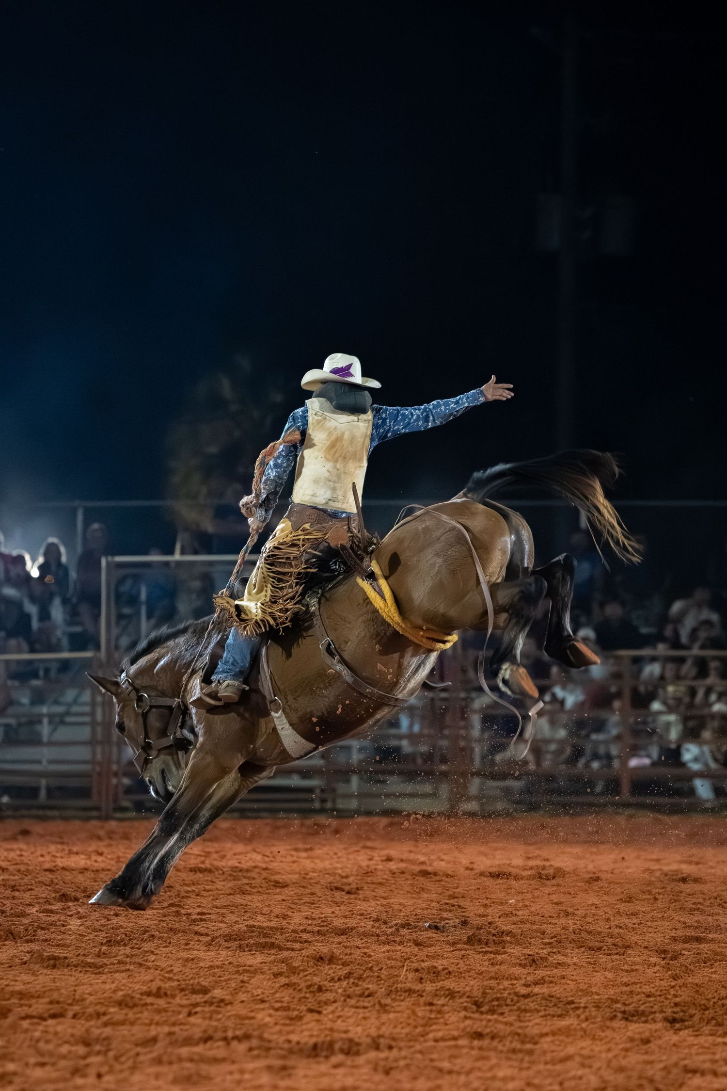 A cowboy rides a bucking bronco at a night rodeo on red dirt arena with crowd watching in background.
