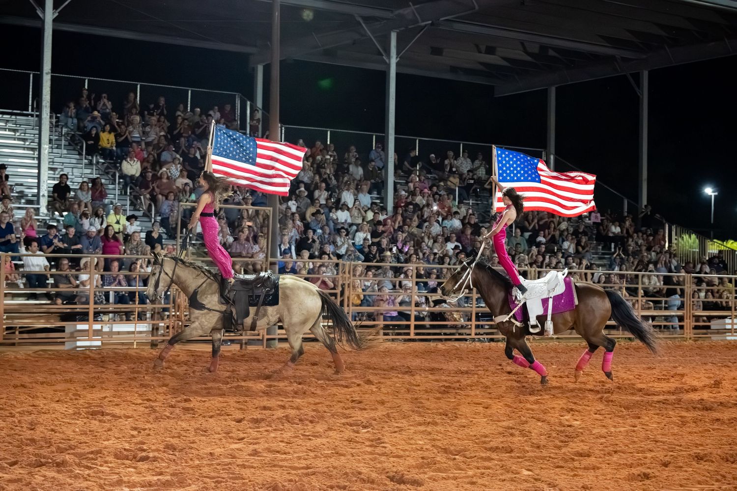 Two riders carrying American flags on horseback at a packed nighttime rodeo arena.