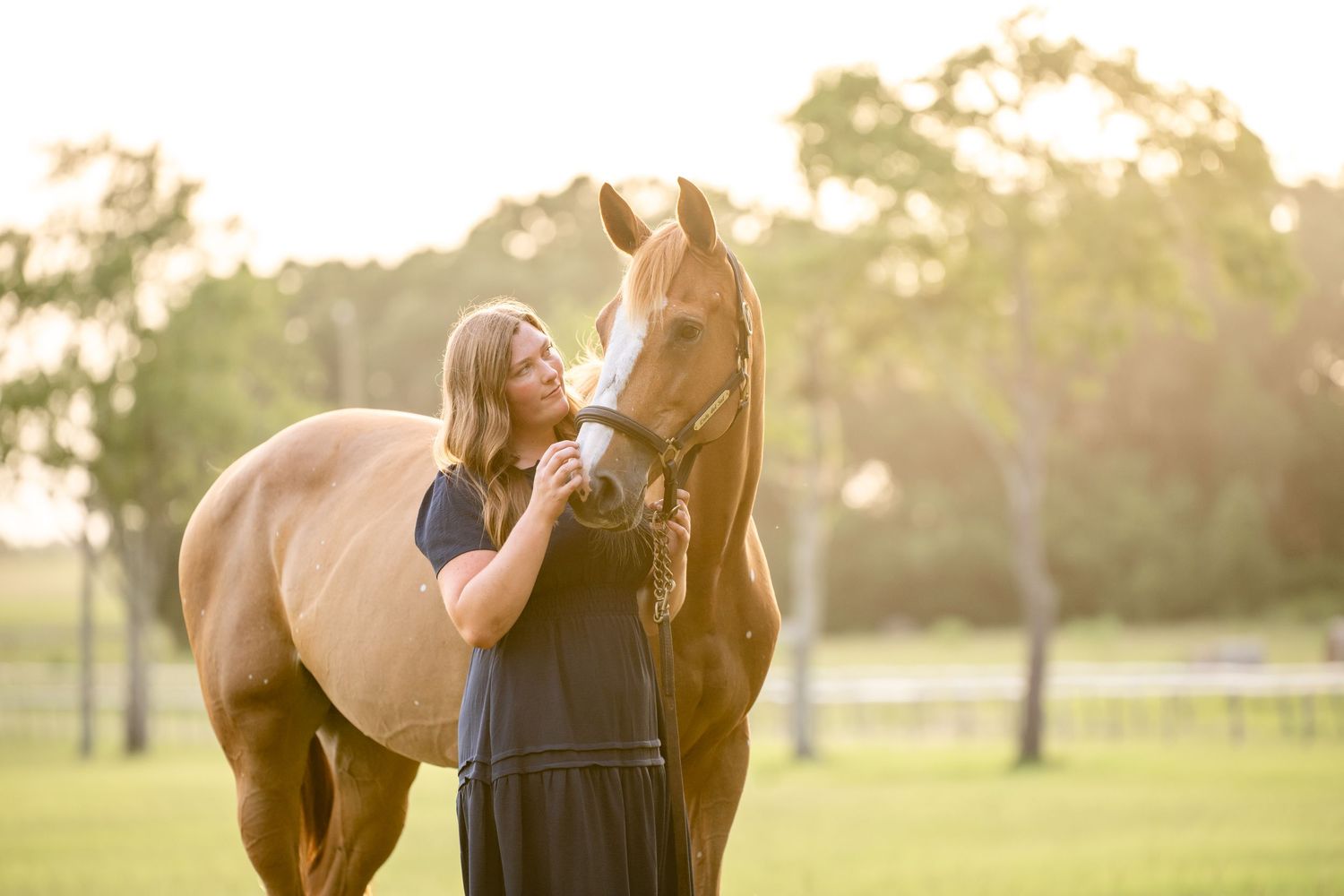 Blonde woman in dark dress shares tender moment with chestnut horse at golden hour.