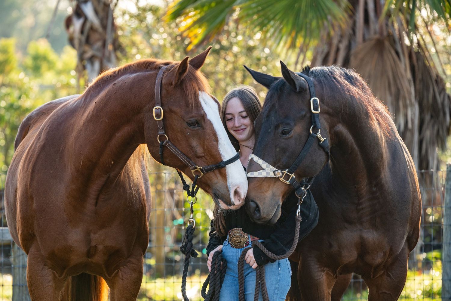 Young woman smiling between two horses, a chestnut and dark bay, outdoors with palm trees.