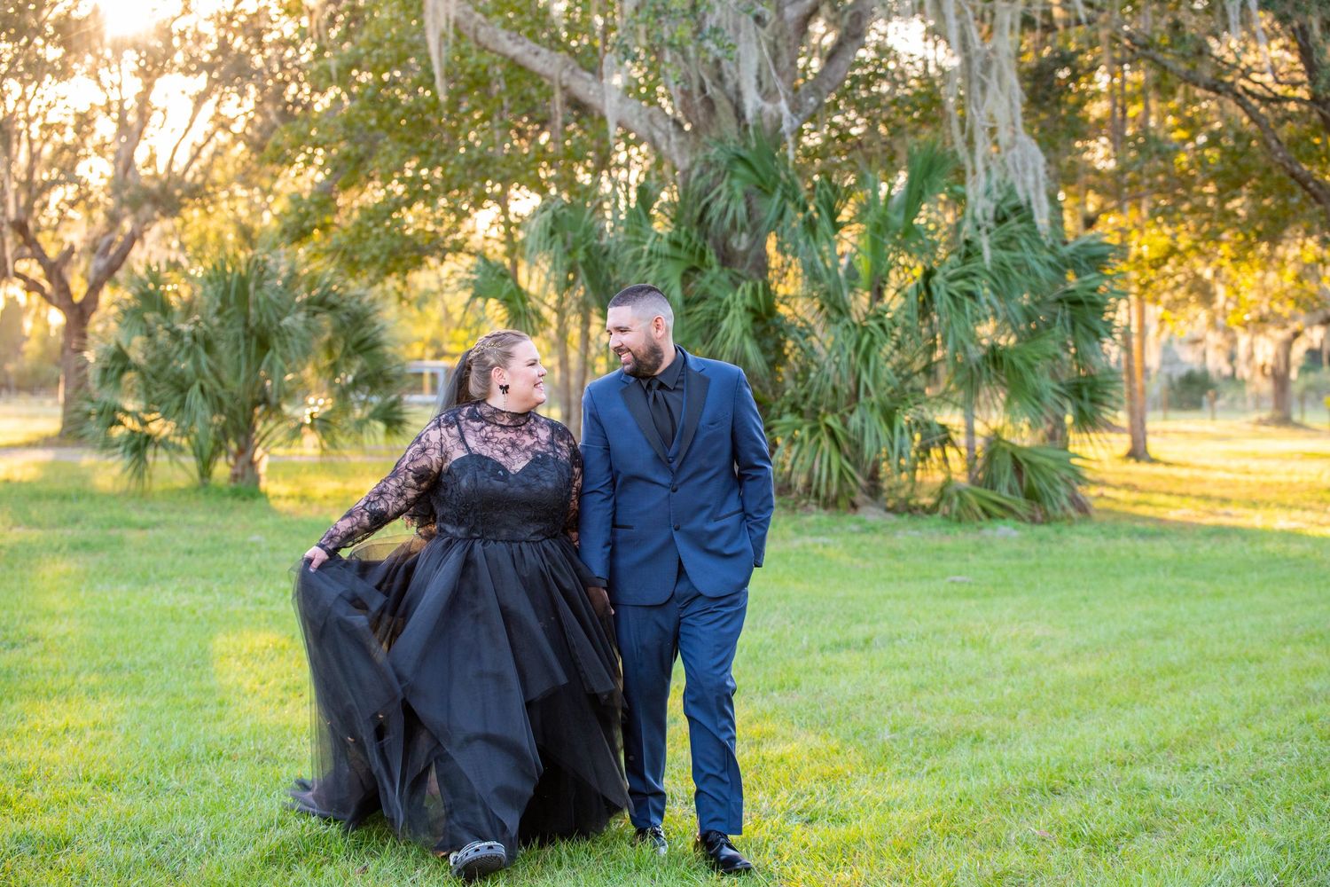 Couple in formal black and blue attire walk hand in hand through a sunlit park at golden hour.